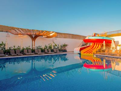 Moderner Hotelpool mit Wasserrutsche und Sonnenliegen unter blauem Himmel.