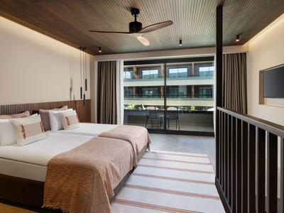 Moderní hotelový pokoj s manželskou postelí, stropním ventilátorem a balkonem s výhledem na sousední budovy.