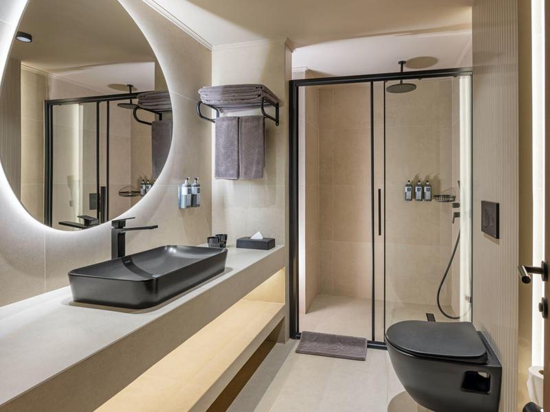 Moderní koupelna s černou baterií, osvětleným kulatým zrcadlem a skleněnou sprchou.