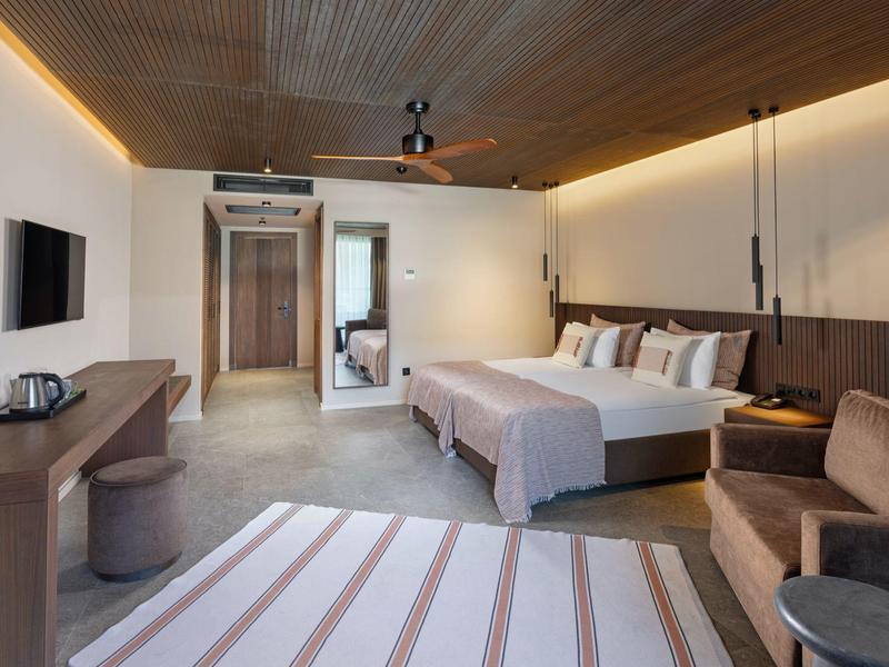 Moderní hotelový pokoj s manželskou postelí, televizí, posezením a koupelnou se skleněnou stěnou.