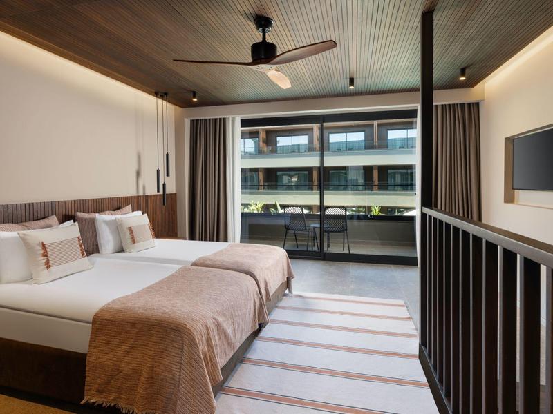 Moderní hotelový pokoj s manželskou postelí, stropním ventilátorem a balkonem s výhledem na sousední budovy.
