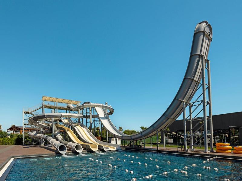 Velká vodní skluzavka s několika dráhami vedle bazénu pod jasně modrou oblohou.