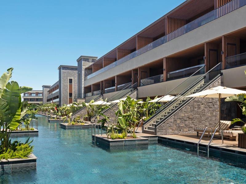 Moderní hotelový komplex s velkým bazénem a tropickými rostlinami pod jasnou oblohou.