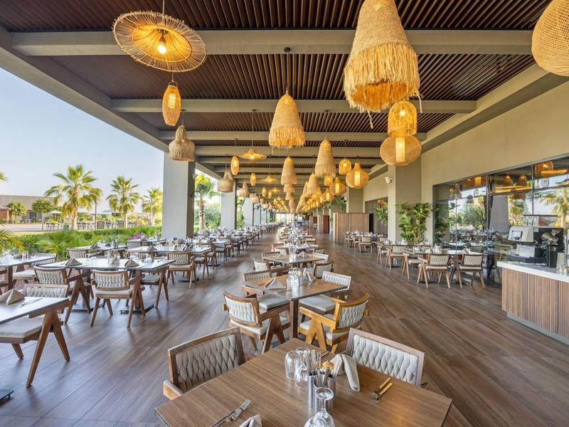 Otevřená restaurace s dřevěnými stoly a židlemi, závěsnými lampami a výhledem na palmy.