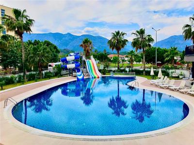 Piscina esterna rotonda con scivoli d'acqua, sdraio e palme davanti a uno sfondo montano.