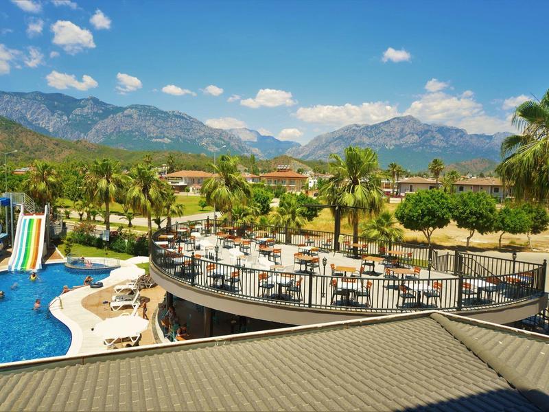 Vista di una piscina con scivoli d'acqua, terrazza soleggiata e palme esotiche davanti a uno sfondo montano.
