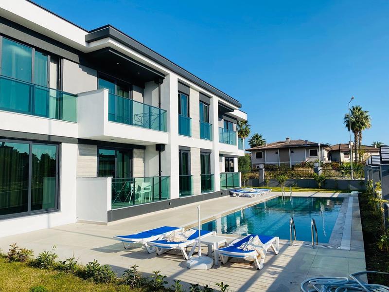 Complesso alberghiero moderno con piscina all'aperto, lettini e vista sul verde sotto un cielo limpido.