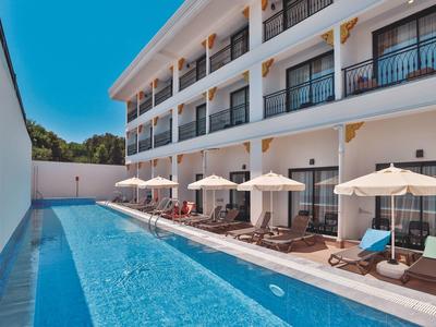 Moderner Hotelpool mit Liegestühlen und Sonnenschirmen neben mehrstöckigem Gebäude bei blauem Himmel.