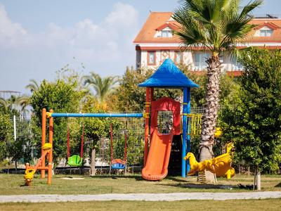 Kinderspielplatz mit Rutsche, Schaukel und Klettergerüst, umgeben von Bäumen und grüner Wiese.