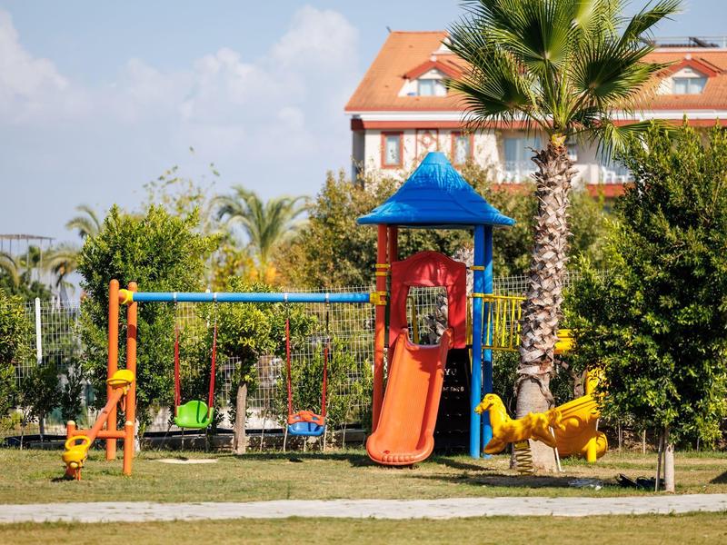 Aire de jeux avec toboggan, balançoire et structure d'escalade devant palmiers et hôtel.