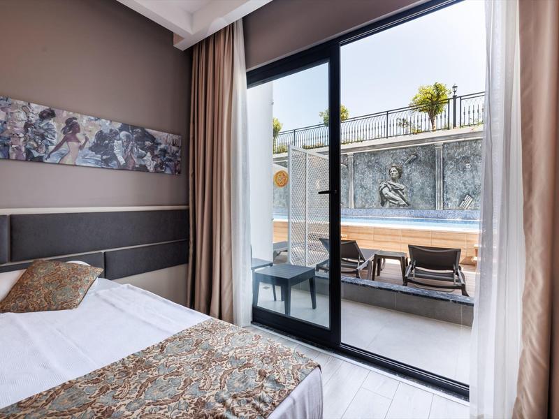 Chambre d'hôtel lumineuse avec lit double et vue sur une terrasse avec piscine et chaises longues.