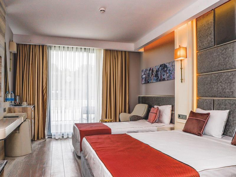 Chambre d'hôtel lumineuse avec deux lits, accents rouges et grande fenêtre avec rideaux.