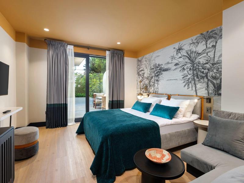 Helles Schlafzimmer mit Holzboden, grüner Bettdecke, grauem Sofa und Naturwandgestaltung.