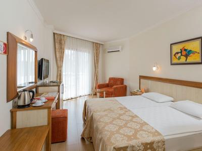 Helles Hotelzimmer mit Doppelbett, Fernseher, Sessel, Schreibtisch und Balkon mit Vorhängen.