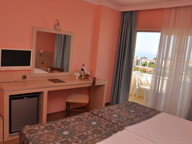 Camera d'hotel con letto matrimoniale, scrivania, sedia, minibar e balcone con tende.