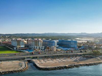 View of a seaside hotel with pool area, promenade, and mountains on the horizon under clear sky.