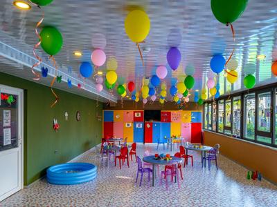 Bunter Kinderspielraum mit Tischen, Stühlen, Luftballons und einem kleinen blauen Pool.