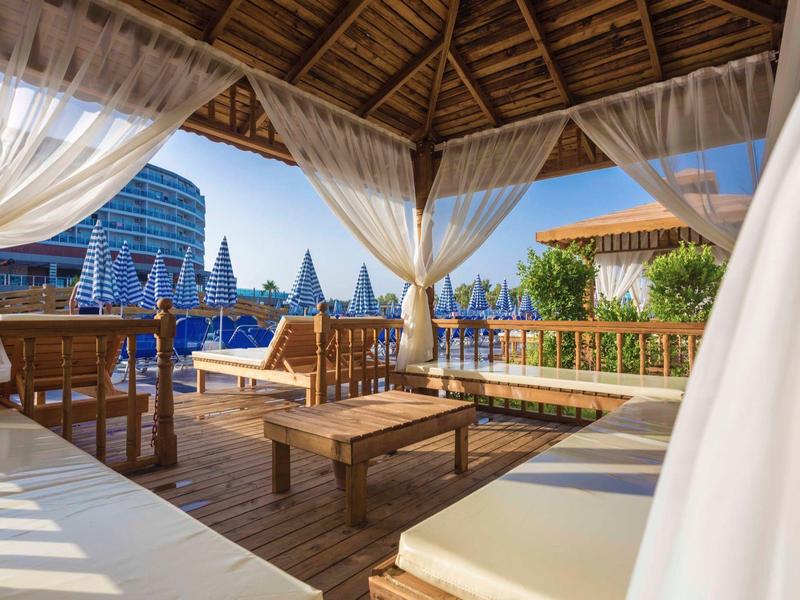 Salon couvert avec mobilier en bois et rideaux blancs près d'une piscine ensoleillée.