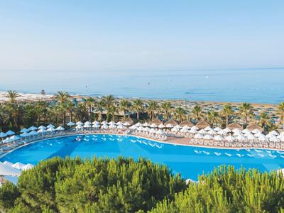 Großer blauer Pool mit weißen Sonnenschirmen und Palmen, dahinter blaues Meer und klarer Himmel.