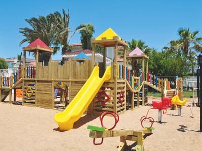 Bunter Spielplatz mit Holzgerüst, Rutschen, Schaukeln und Sandboden unter blauem Himmel.