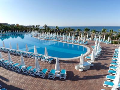 Großer, ovaler Pool mit vielen blauen Liegestühlen und weißen Sonnenschirmen am Meerufer.