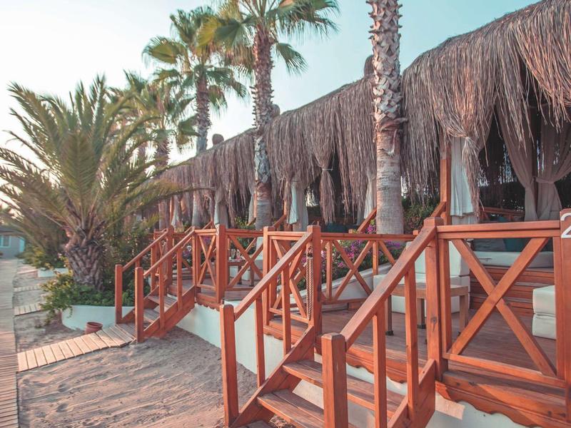 Houten trappen leiden naar hutten aan het strand met palmbomen en een heldere lucht.