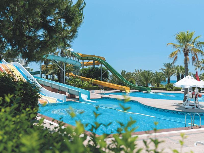 Buitenzwembad met glijbanen, omgeven door bomen en palmbomen onder een heldere blauwe lucht.