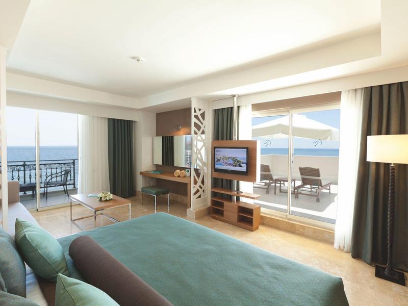 Lichte hotelkamer met groot bed, tv, balkon met tafel en zeezicht.