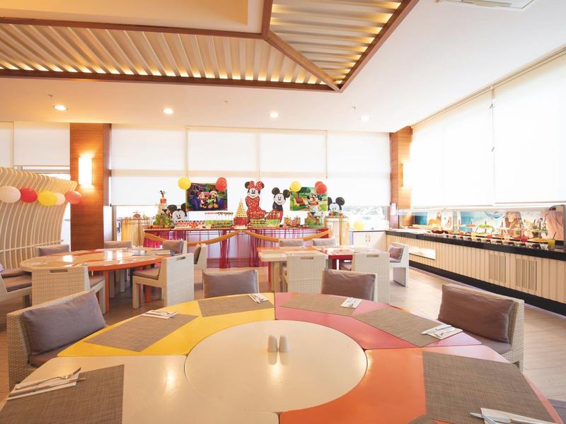 Lichte eetzaal met kleurrijke tafels en stoelen naast grote ramen in een hotelrestaurant.