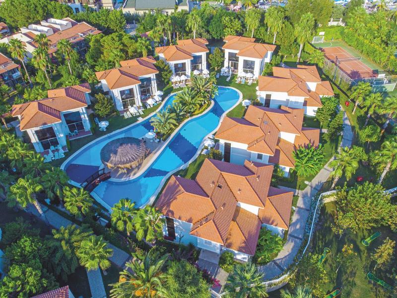 Luchtfoto van een vakantieresort met meerdere gebouwen en een gebogen zwembad te midden van groen.