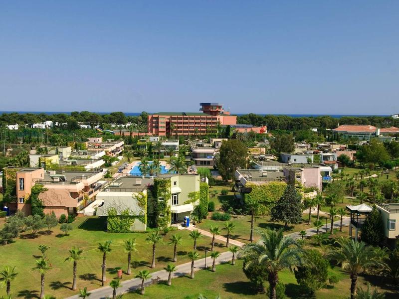 Pohled na hotelový resort se zelenými zahradami, bazény a vícepatrovými budovami pod jasnou oblohou.