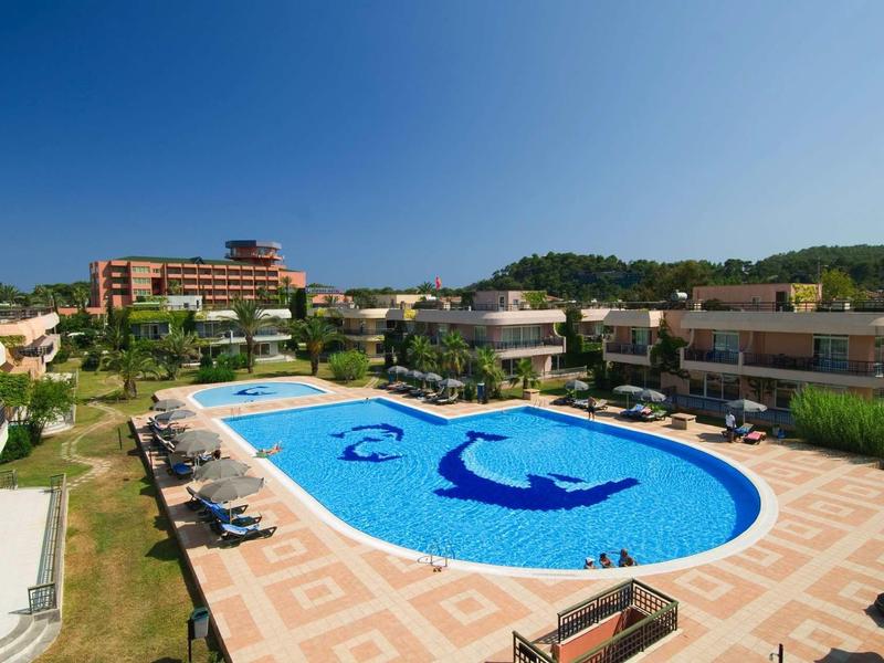 Hotel s velkým bazénem obklopeným lehátky a budovami pod jasnou modrou oblohou.