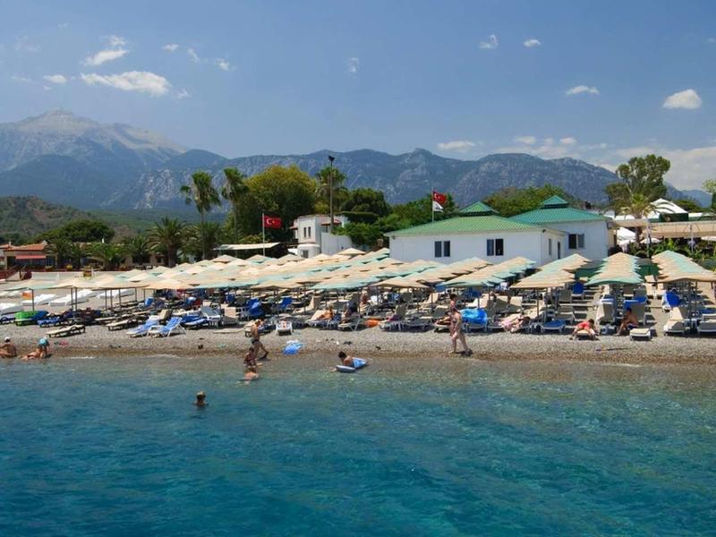 Pláž s čistou modrou vodou, slunečními parasoly a horami v pozadí.