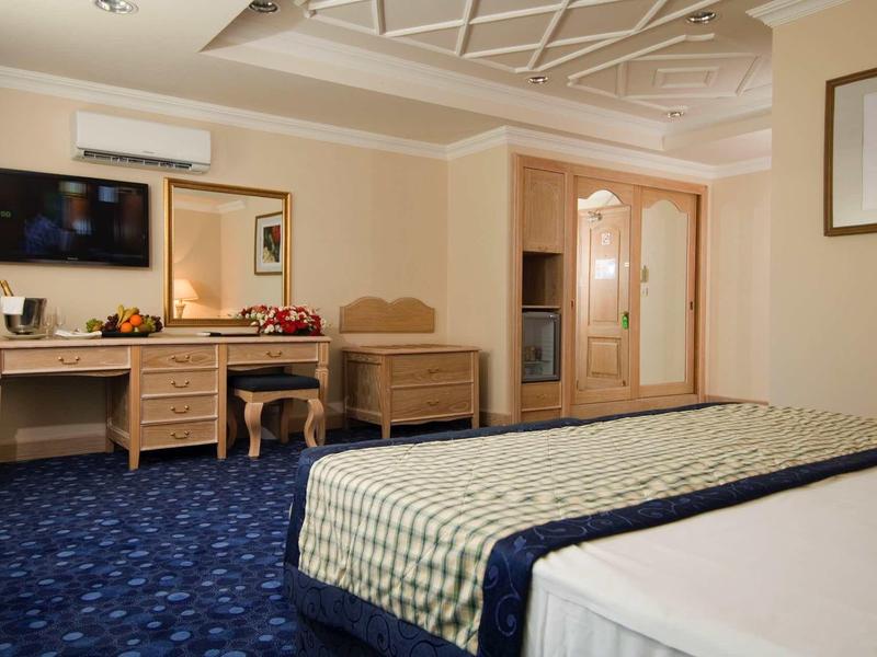 Hotelový pokoj s modrým kobercem, manželskou postelí, stolem, židlí a televizí na zdi.