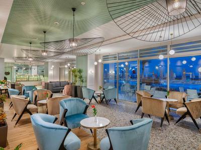 Area lounge moderna in hotel con poltrone comode, tavoli e grandi finestre con vista.