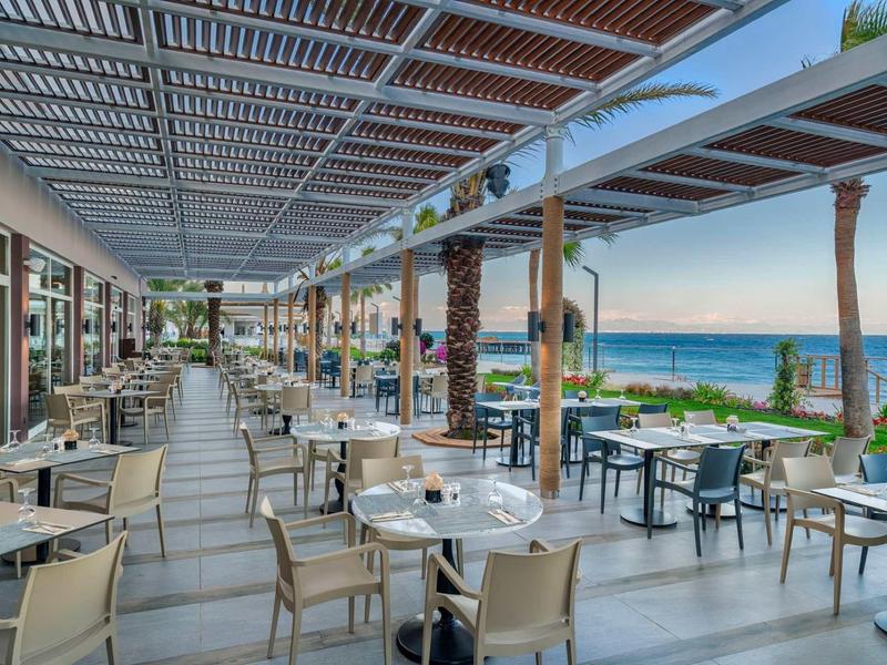 Nowoczesny taras restauracyjny z widokiem na morze i palmy, jasne meble i przestronne wnętrze.