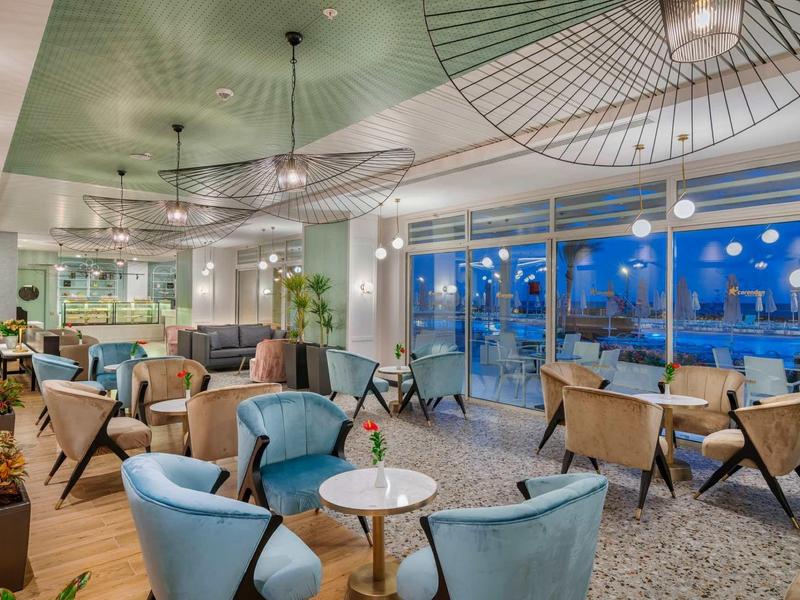 Nowoczesny salon hotelowy z niebieskimi fotelami, okrągłymi stolikami i dużymi, jasnymi oknami.