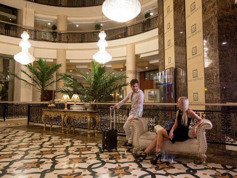 Dvě osoby s zavazadly sedí a stojí v elegantní hotelové hale s mramorovou podlahou a velkými rostlinami.