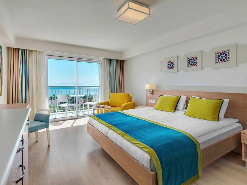 Lichte hotelkamer met groot bed, blauwe en groene accenten, balkon en zeezicht.