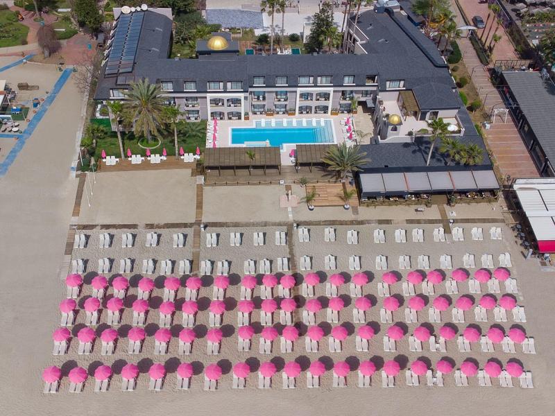 Vista aerea di un hotel con piscina e lettini da spiaggia con ombrelloni rosa sulla sabbia.