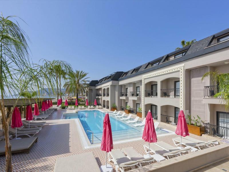 Lobby dell'hotel moderna con piscina, lettini e ombrelloni rossi sotto un cielo terso.