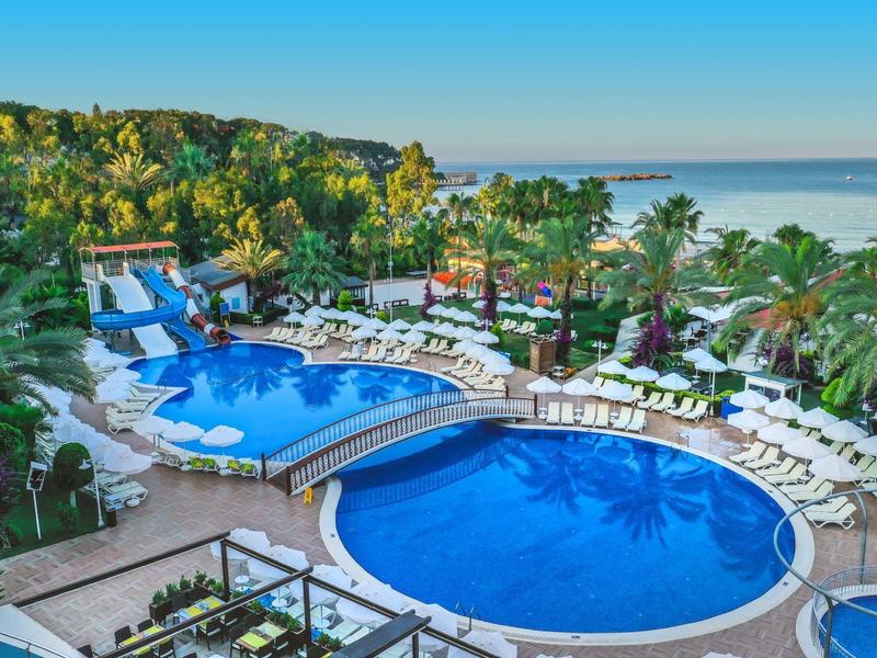 Hotel resort with two swimming pools, surrounded by palm trees and sun loungers, by the sea.