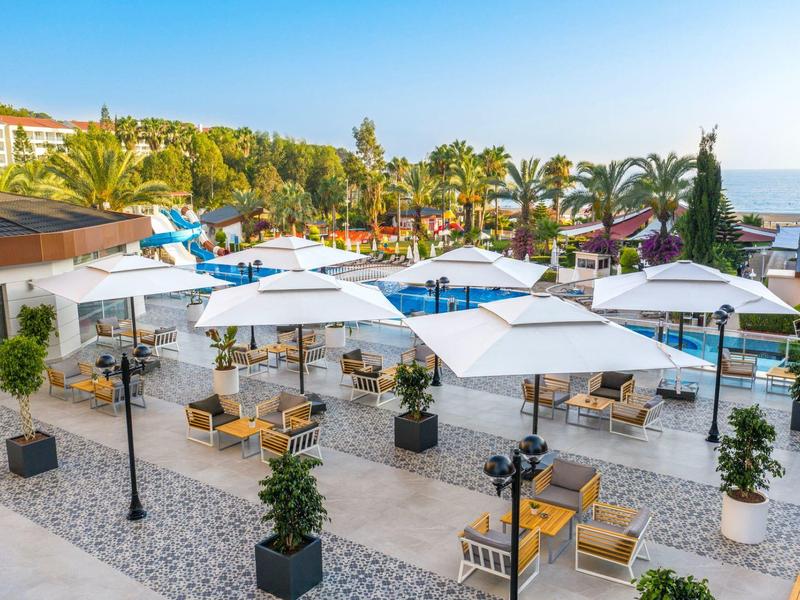 Outdoor patio area with umbrellas, tables, and chairs in a sunny hotel garden next to the beach.