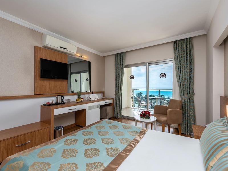 Bright hotel room with a large bed, desk, TV, and balcony overlooking the sea.