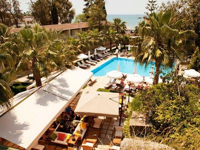 Hotelpool mit blauer Wasserfläche, Liegestühlen, Palmen und schattigen Sitzbereichen im Freien.