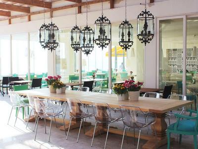 Jasna, nowoczesna jadalnia z dużymi drewnianymi stołami, kolorowymi krzesłami i dekoracyjnymi żyrandolami.