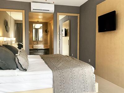 Nowoczesny pokój hotelowy z łóżkiem podwójnym, szarym narzutą i telewizorem na ścianie.