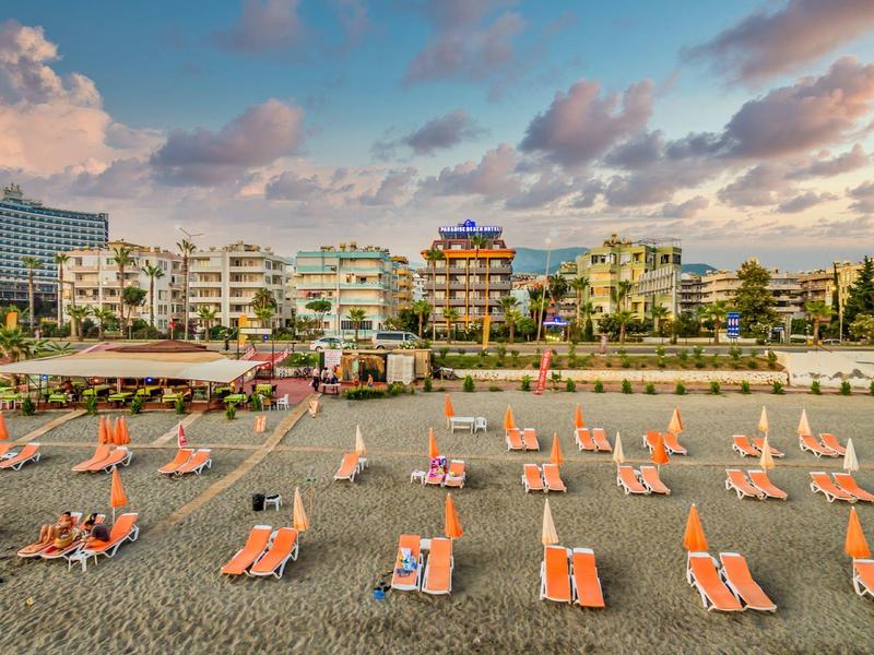 Beach with orange sunbeds and umbrellas in front of a hotel complex under a cloudy sky.