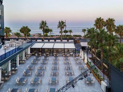 Terrasse mit zahlreichen Tischen und Stühlen, umgeben von Palmen und Blick auf das Meer.