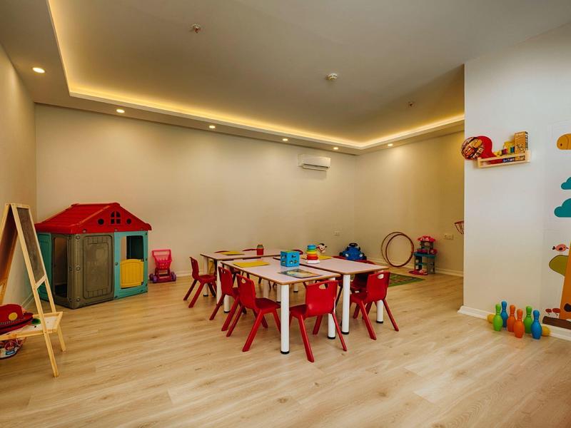 Sala giochi per bambini con tavoli, sedie rosse e giocattoli colorati.